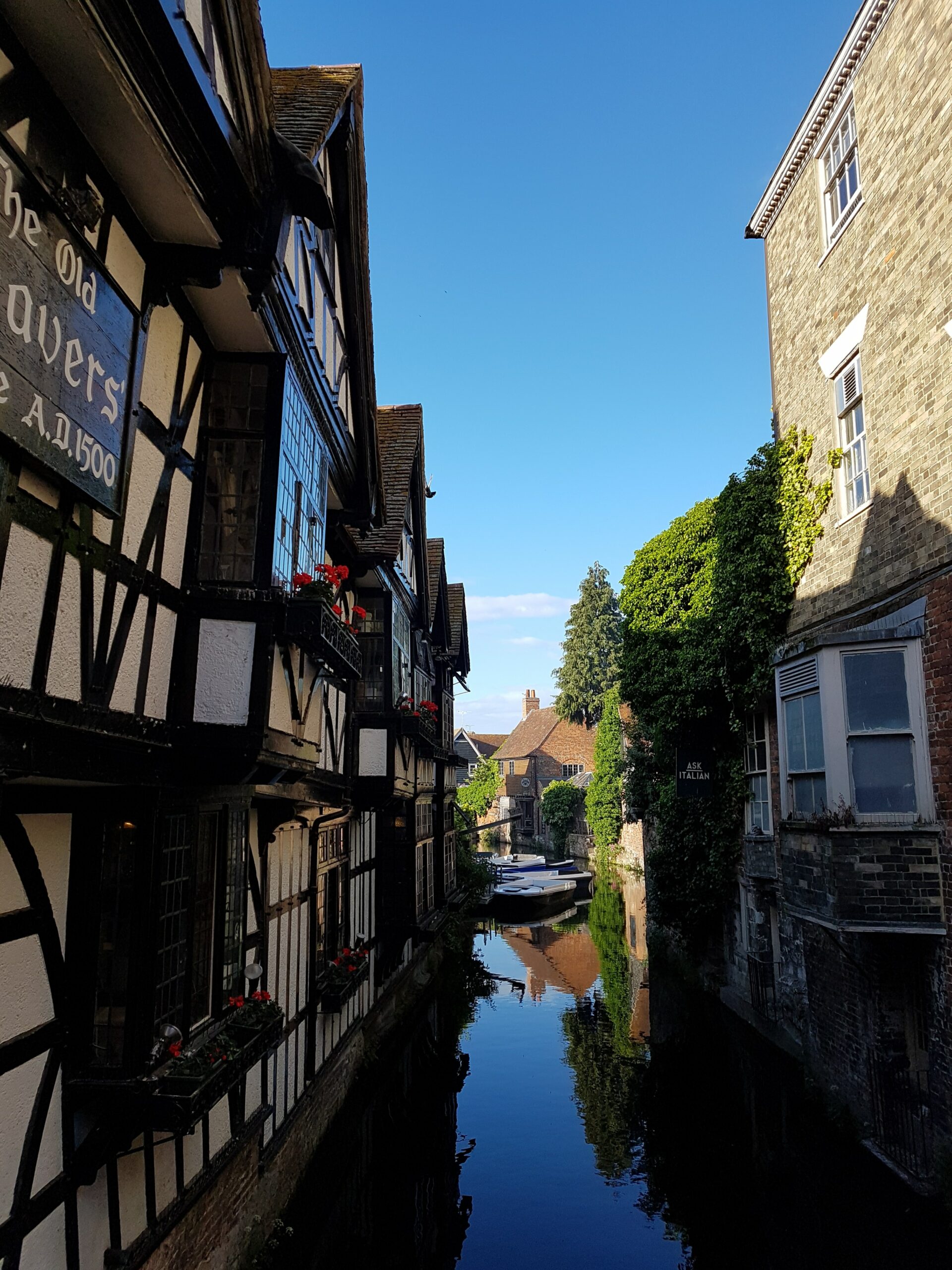 A Tudor house on the left and a older style property on the right with a river stream in the middle on a clear, sunny day.